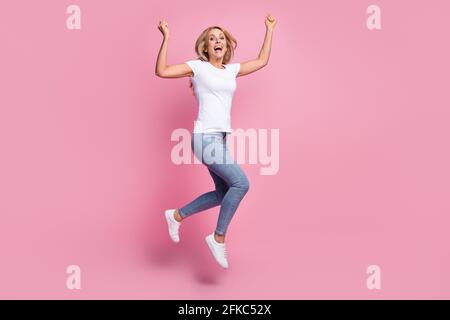 Photo of crazy astonished lady jump raise fists wear white t-shirt jeans footwear isolated pink color background Stock Photo