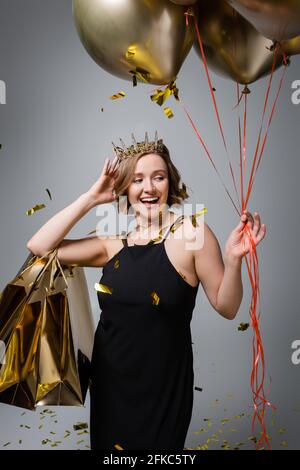 happy plus size woman in slip dress and crown holding golden balloons and shopping bags near confetti on grey Stock Photo