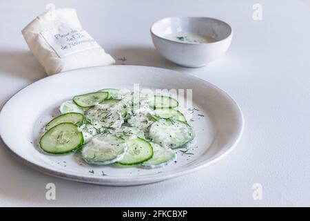 Plate of creamy yogurt cucumber salad with a bowl of yogurt dressing and a bag of fleur de sel in the background. Stock Photo