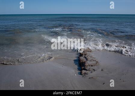 The Mediterranean coast with sand against a blue sky with a ruined jetty made of old stones, some of which protrudes from the waves Stock Photo