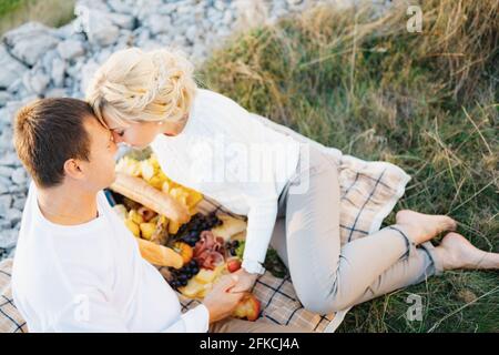 Barefoot woman leaned her forehead against man sitting on a blanket. Man and woman are holding hands. There is food on the blanket Stock Photo