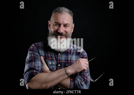 Studio portrait of mature bearded man in casual clothes with eyeglasses in hand smiles and looks directly at camera, informal lifestyle, black backdro Stock Photo