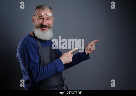 Follow me. Look here. Close up portrait of cheerful pleased qualified experienced joyful apron-wearing grandfather pointing on empty blank place over Stock Photo