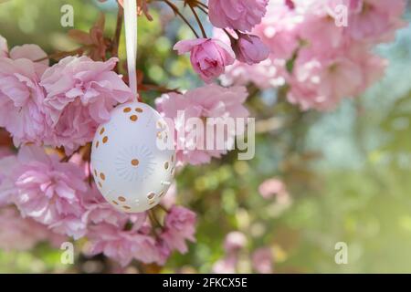 Decorative Easter egg hanging on flowering cherry tree with pink flowers. Easter background. Blossoms on a sunny spring day. Selective focus Stock Photo