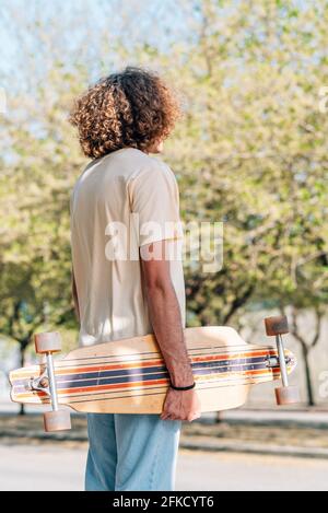 vertical take from the back of a caucasian man holding a skateboard in the street. He has curly long hair and dresses casual clothes Stock Photo