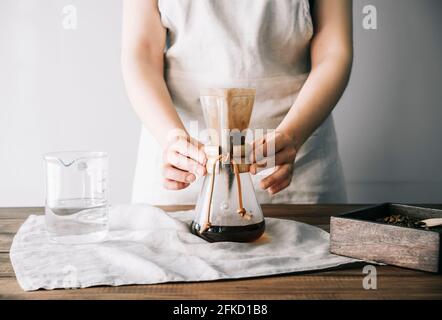 Person in apron holding manual glass coffee pot with filter and fresh coffee on a dark wooden table Stock Photo