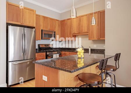 An apartment kitchen with maple cabinets, black granite countertops, stainless appliances and pendant lights, and bar stools. Stock Photo