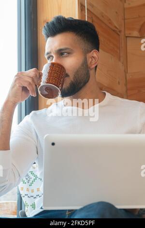 Indian man wearing a white shirt and sipping coffee sitting next to the window with his laptop. Looking outside through the window. Cozy room. Winter season in the city of Zakopane, Poland.  Stock Photo