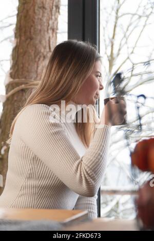 Attractive young woman looking outside through a glass window. Holding coffee mug. The tree trunk can be seen through the window in the background. High-quality photo. Zakopane, Poland.  Stock Photo