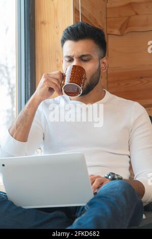 Focused young man sipping coffee and working on his laptop. Sitting next to the window in a peaceful cozy room. Wearing a white shirt and jeans. Winter season in the city of Zakopane, Poland.  Stock Photo
