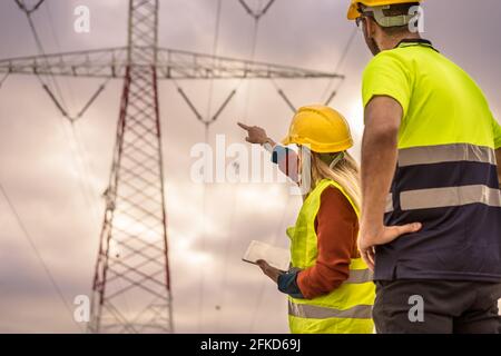 Picture of two electrical engineers using a tablet standing at a High-voltage tower to view the planning work by producing electrical energy at high v Stock Photo