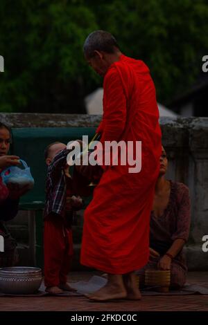 Luang Prabang, Laos - July 6, 2016:  A Buddhist monk accepts rice given to him by a child during the early morning alms giving ceremony of Sai Bat. Stock Photo