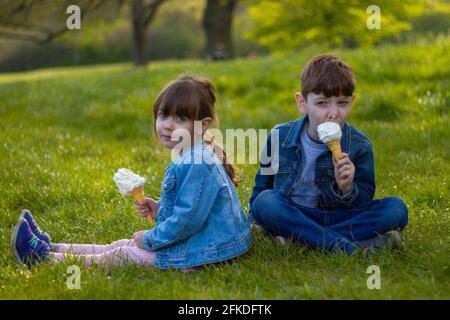 Portrait of a cute boy and a girl wearing blue jackets sitting on a lawn on a sunny day eating ice cream Stock Photo