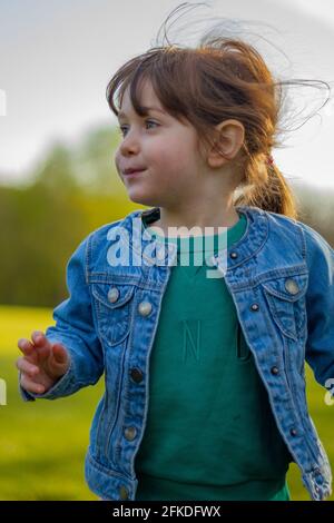 Portrait of a cute, brown-haired, blue-eyed baby girl wearing a blue jacket and green jumper running in a park on a sunny day Stock Photo