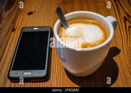 White coffee cup and cell phone on wooden table. Stock Photo