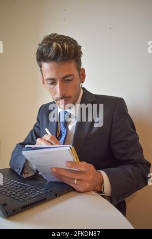 Vertical shot of a Spanish Caucasian businessman writing on his calendar and using his laptop Stock Photo