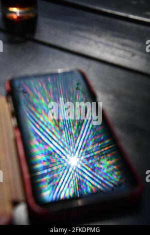Bright rainbow rays reflect in mobile phone screen surface with blurred background. Multicolored beams and reflection atop smartphone screen closeup Stock Photo
