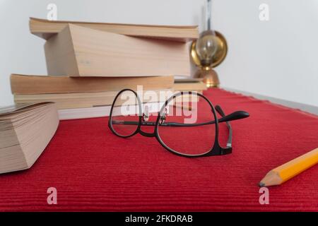 Eyeglasses in foreground, a pencil on the right and a book stack and an oil lamp in the background, lying on a red tablecloth. High quality photo Stock Photo