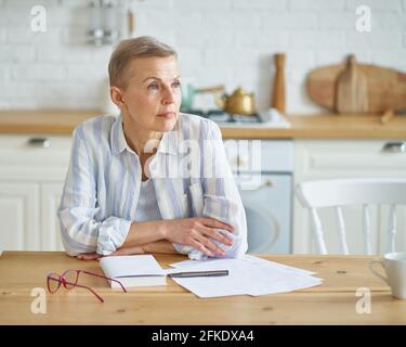 Beautiful mature woman looking aside and thinking about something while sitting at wooden table in kitchen and working with documents. Senior people and distance work concept Stock Photo