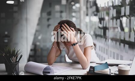 Businesswoman sitting in her workplace and holding head with hands feeling desperate or suffers from headache. Stock Photo
