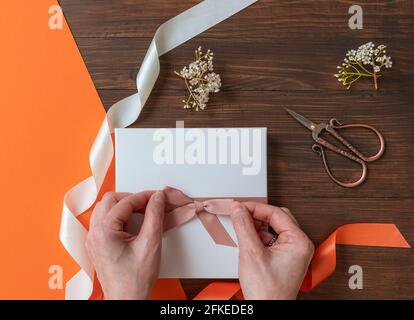 https://l450v.alamy.com/450v/2fkede8/female-hands-make-a-pink-bow-on-a-white-box-placed-on-a-wooden-table-with-flowers-and-colorful-ribbons-2fkede8.jpg