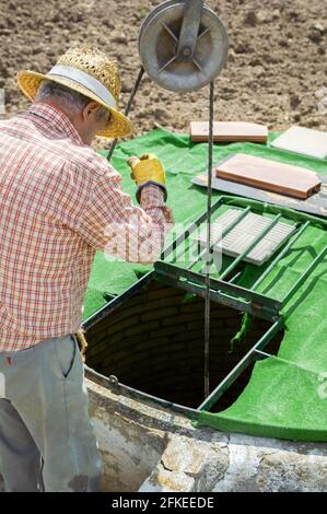 Old man drawing water from a rustic well with pulley in the countryside on a sunny day. Stock Photo