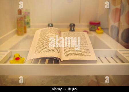 Book resting on bath tray with duck, various bottles and accessories in background seen through steam Stock Photo
