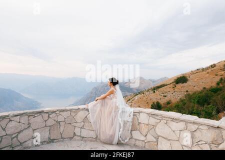 The bride sits on the stone side of the observation deck on Lovchen mountain and looks at the Bay of Kotor  Stock Photo