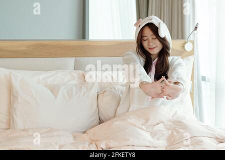 Happy pretty young Asian woman in pajamas sitting on bed and tsretching after waking up early in the morning Stock Photo