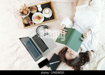 Young woman lying on bed next to laptop and tray with breakfast and reading interesting book, view from the top Stock Photo