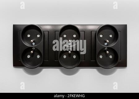 Black frame of a double grounded electrical socket in three rows on the gray wall of the room. Stock Photo