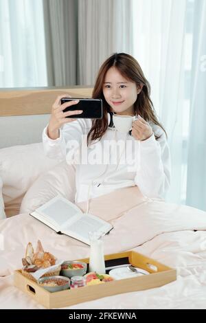 Lovely young Asian woman having breakfast in bed, she is taking selfie or filming video with cup of coffee in hand Stock Photo