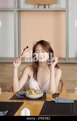 Portrait of pretty smiling young woman closing eyes when eating bowl of delicious poke dish or salad Stock Photo