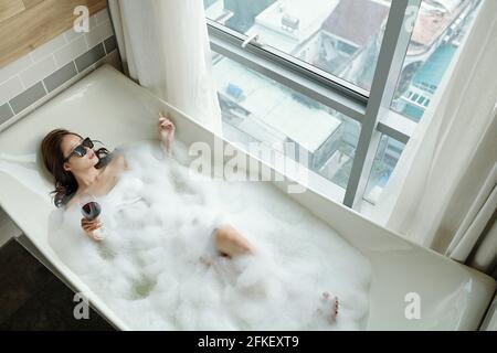 Pretty young woman in sunglasses relaxing in bubble bath with glass of wine, view from above Stock Photo