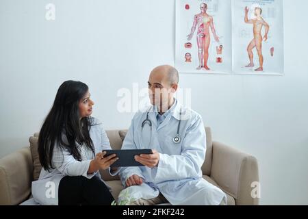 General practitioners sitting on sofa in doctors lounge room and discussing list of patients symptoms on tablet computer Stock Photo