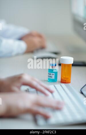Vial of vaccine against coronavirus and bottle of pills on desk in front of doctor working on laptop Stock Photo