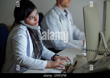 Serious young female Indian doctor talking on phone with patient and entering his complaints and symptoms in form on computer scren Stock Photo