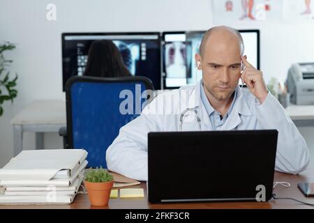 Serious concentrated general practitioner wearing earphones when watching webinar on laptop, his colleague is working in background Stock Photo