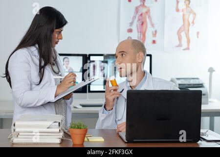 Physician recommending bottle of new pills to his collegue who is taking notes in document on clipboard Stock Photo