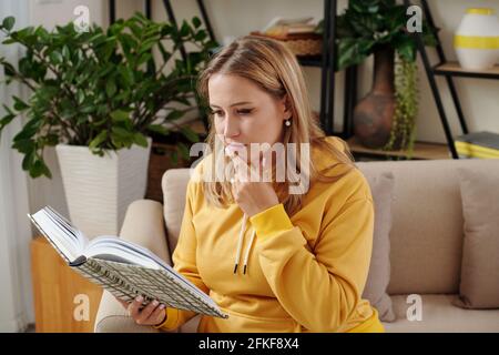 Pensive young woman sitting on sofa at home and reading interesting novel or students book when getting ready for exam Stock Photo