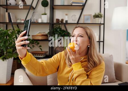 Pretty positive young blond woman biting unpeeled orange and taking selfie for social media Stock Photo