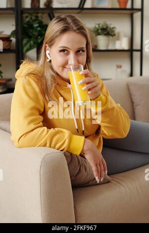 Pretty smiling young woman resting on sofa, listening to audiobook and drinking glass of orange juice Stock Photo