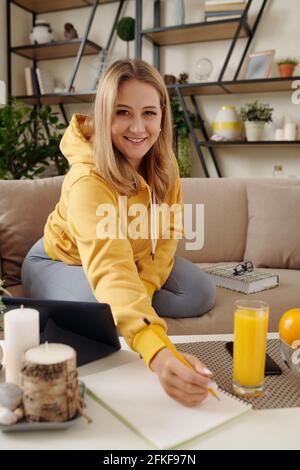 Pretty smiling young woman studying, watching webinar on tablet at home and taking notes in textbook Stock Photo