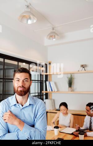 Portrait of handsome confident bearded entrepreneur standing at table, two of his colleagues discussing documents in background Stock Photo