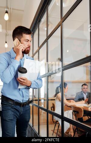 Serious young bearded entrepreneur standing outside the meeting room with glass walls and talking on phone with colleagues Stock Photo