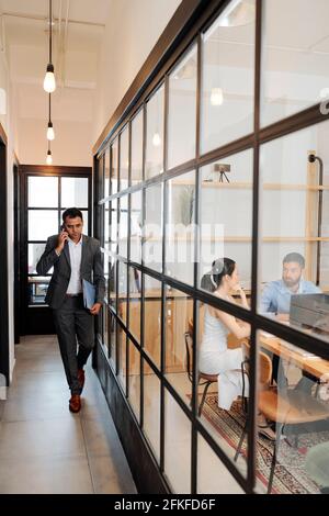 Confident successful Indian entrepreneur walking along office corridor and talking on phone with business partner Stock Photo