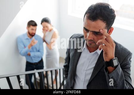 Businessman tired of hearing gossips behind his back, he is walking up the stairs and eavesdropping on conversation of colleagues Stock Photo