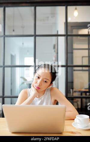 Pensive young Asian businessswoman watching presentation or reading report on laptop screen when sitting at coffee table with cup of coffee Stock Photo