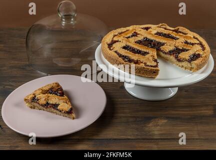 A slice of cherry tart near a white cake stand with the rest of the tart and the glass lid on wooden table Stock Photo