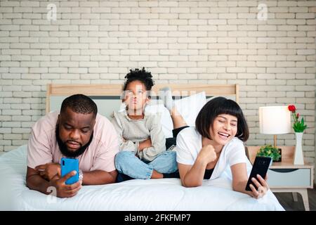 African American girls are not satisfied and sad, sitting in the middle between father and mother. Parents Are using the smartphone and abandoned chil Stock Photo